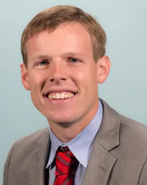 Portrait of a young man wearing a gray suit, blue shirt, and red striped tie against a light blue background.