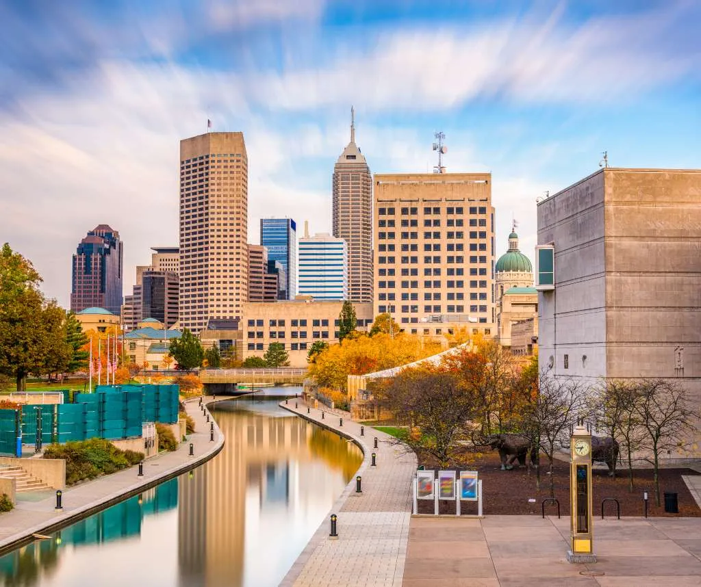 Urban cityscape with modern skyscrapers, a river canal, and autumn foliage under a partly cloudy sky.